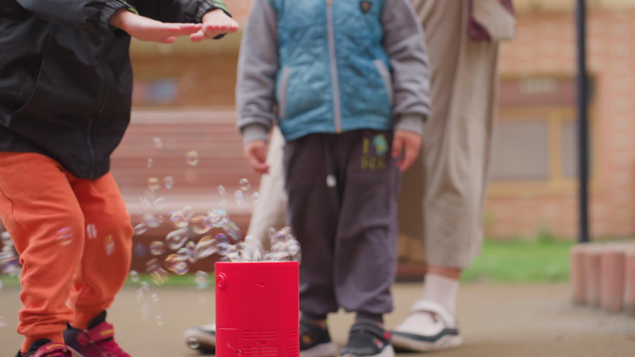 Excited boy in orange trousers bends hitting soap bubbles floating from red bubble machine while another child stands beside watching closely, playful outdoor scene filled with curiosity