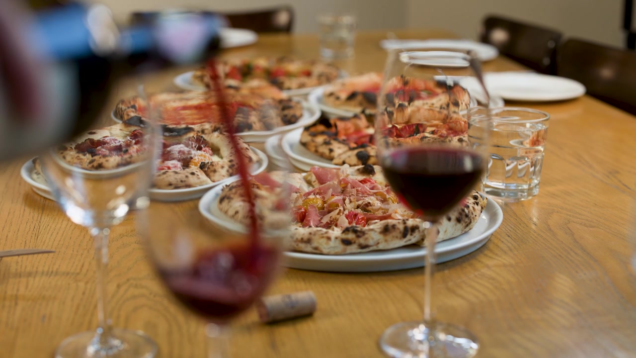 Vino rosso che si versa in un bicchiere su un tavolo di legno con pizze napoletane e prosciutto