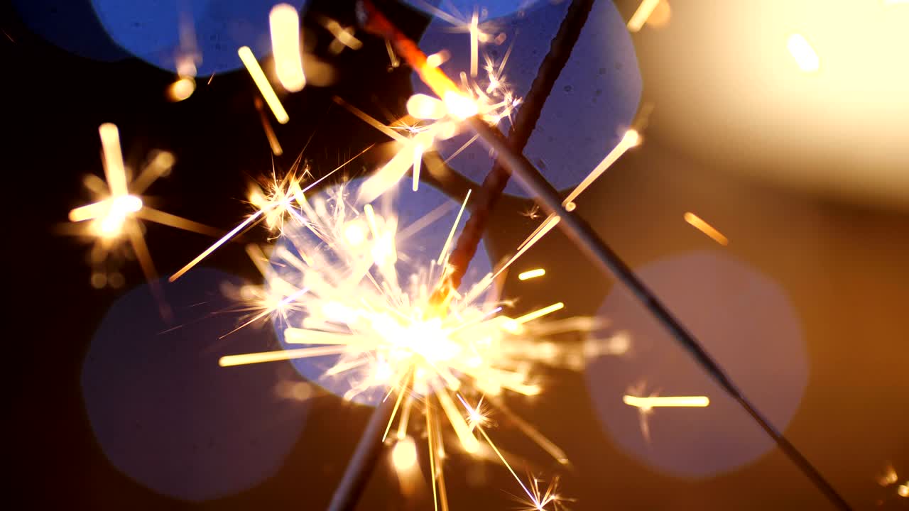 una toma macro en primer plano de dos chispas de año nuevo en llamas cruzadas. chispas bengal dispersándose en pequeñas chispas contra un fondo de guirnaldas azules en borrón. cámara lenta 4k