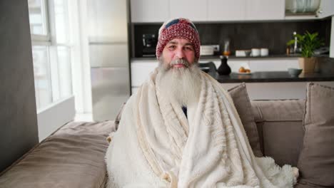 Portrait-of-a-smiling-elderly-man-with-a-lush-gray-beard-who-is-wrapped-in-a-warm-white-blanket-and-sits-in-a-hat-on-a-brown-sofa-in-a-modern-apartment