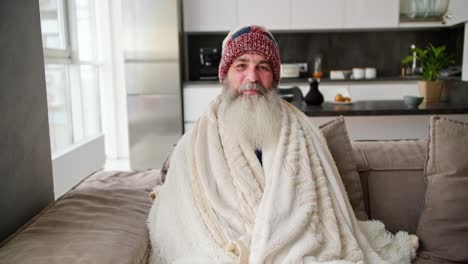 retrato de un anciano sonriente con una barba gris exuberante que está envuelto en una manta blanca cálida y se sienta en un sombrero en un sofá marrón en un apartamento moderno
