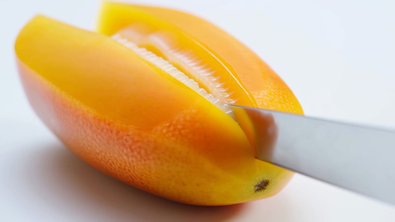 Un coltello in acciaio inossidabile taglia mezze manghi maturi, separando la polpa lucida giallo-arancio e rivelando una consistenza umida e fibrosa in un'illuminazione da studio pulita e minimale con uno styling alimentare luminoso e preciso