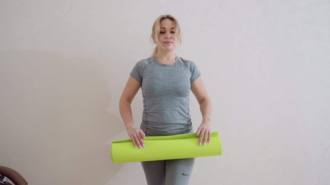 Confident sportswoman in fitted grey workout clothes spreads bright green fitness mat while standing indoors in neutral room, preparing for home exercise