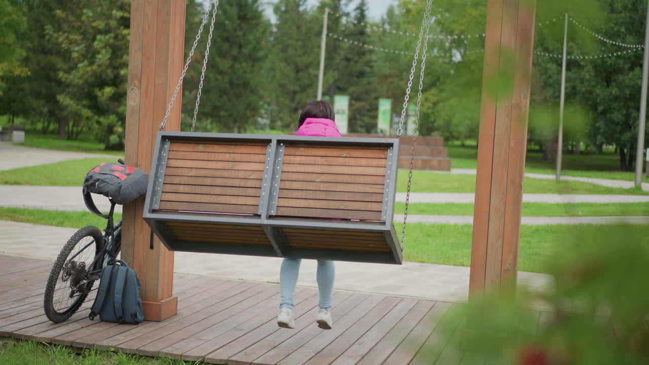 Žena v růžové bundě na houpačce, dřevěná lavice zavěšená na pergole vedle zaparkovaného kola a batohu, rozmazané listí v popředí, zatažená obloha, klidná promenáda v parku, zamyšlená póza, zpomalený záběr