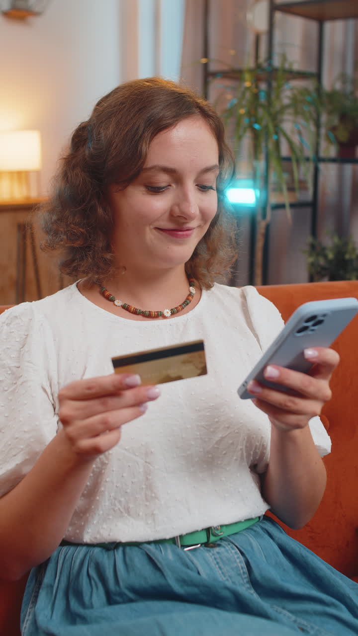 Giovane donna felice che usa la carta di credito e lo smartphone mentre fa acquisti online seduta sul divano