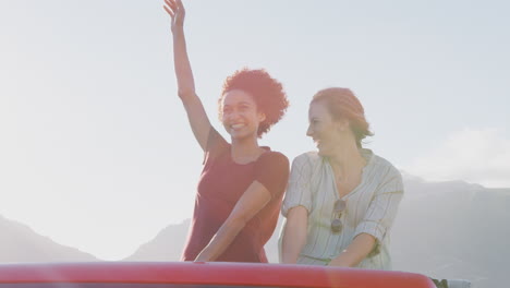 two female friends standing up in back of open top hire car on summer vacation