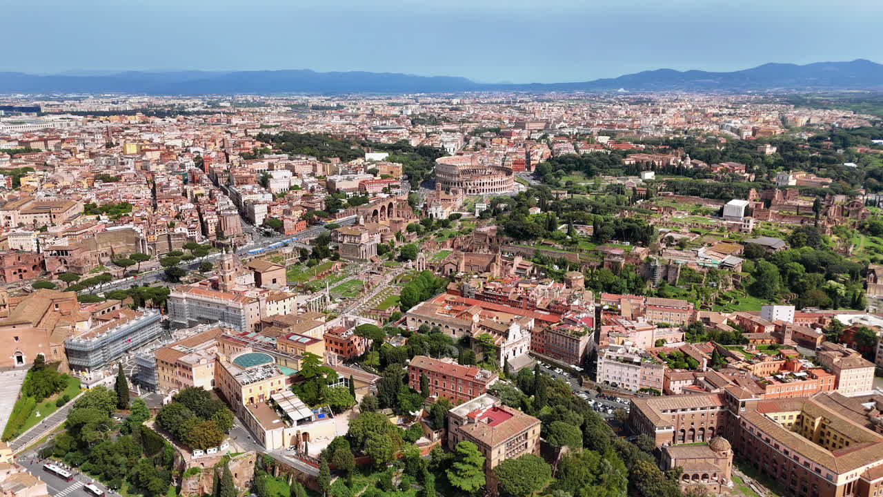 Aerial drone view of Rome's historic center featuring the Colosseum, Roman Forum, and Palatine Hill. Ancient ruins surrounded by modern cityscape under clear daylight