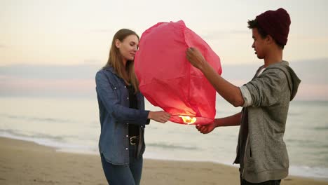 Junges-Multiethnisches-Paar-Hält-Vor-Dem-Start-Eine-Rote-Papierlaterne-In-Der-Hand.-Romantisches-Date-Am-Strand.-Attraktive-Frau-Zusammen-Mit