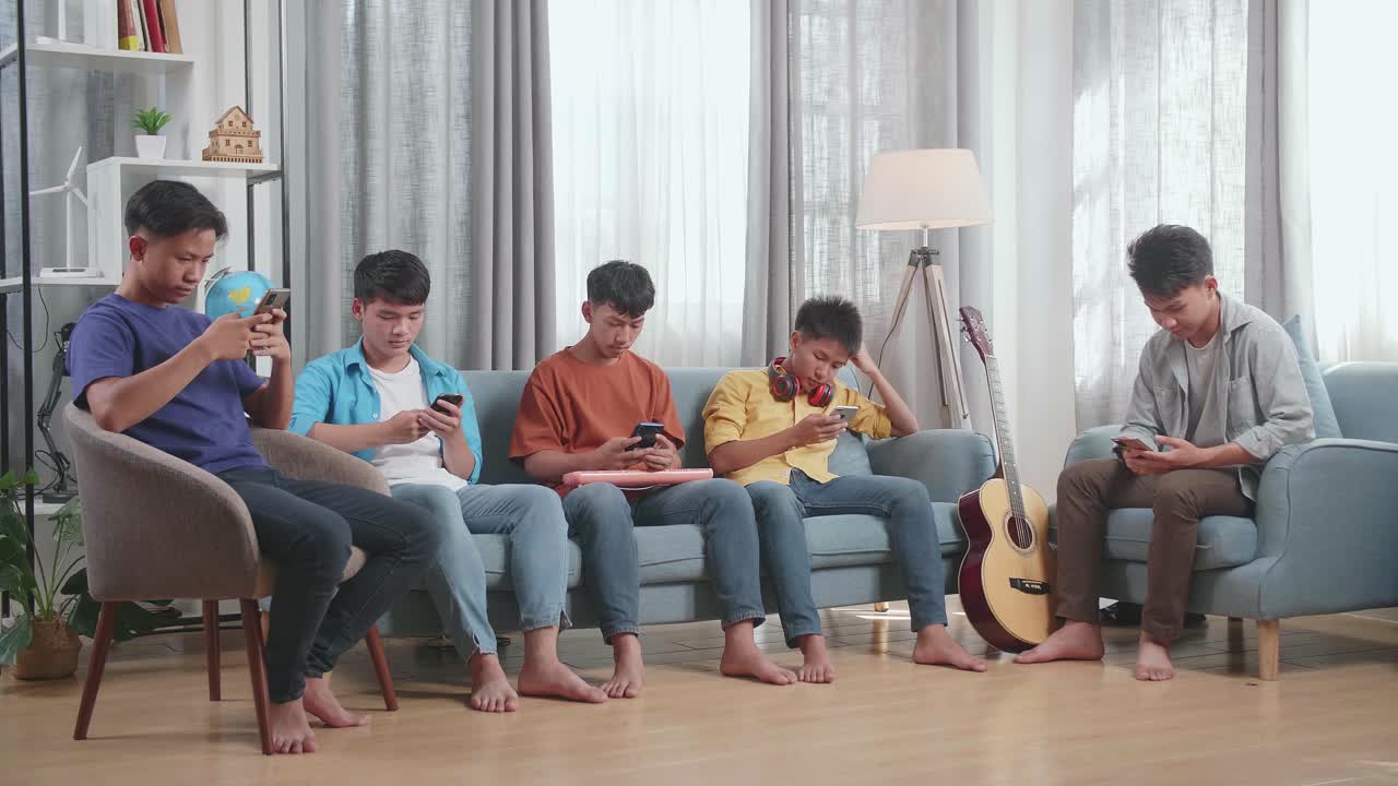 음악 악기 를 들고 있는 젊은 아시아인 친구 들 의 그룹 은 집 의 소파 에 앉아 스마트폰 을 연주 하는 것 을 즐긴다