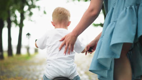 back view of a woman with painted nails supporting her son as he learns to ride a bicycle, the young boy uses his leg to balance himself as they move forward on a path