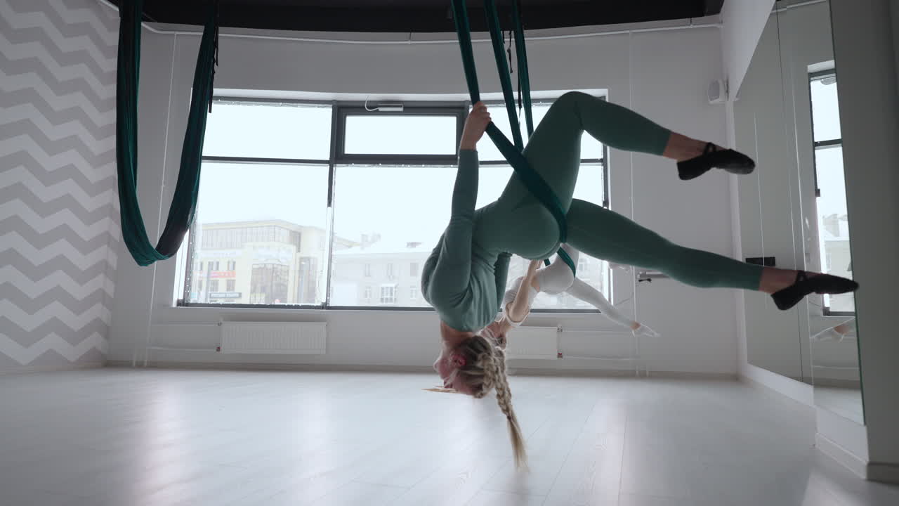 Two young yogi women doing aerial yoga practice in green hammocks in fitness club. Beautiful females working out in class performing aero yoga. Variation of Parsvottanasana Pyramid pose