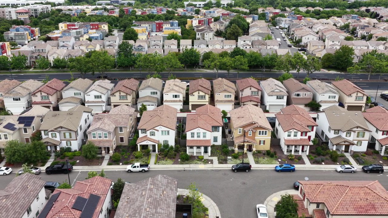 Rumah-rumah pinggiran kota dua lantai dengan atap terakota mengapit jalan raya lebar, rumah-rumah kota berwarna-warni dengan panel surya dan mobil-mobil yang diparkir di kedua sisi di bawah langit cerah. California, AS. Pandangan udara dari atas