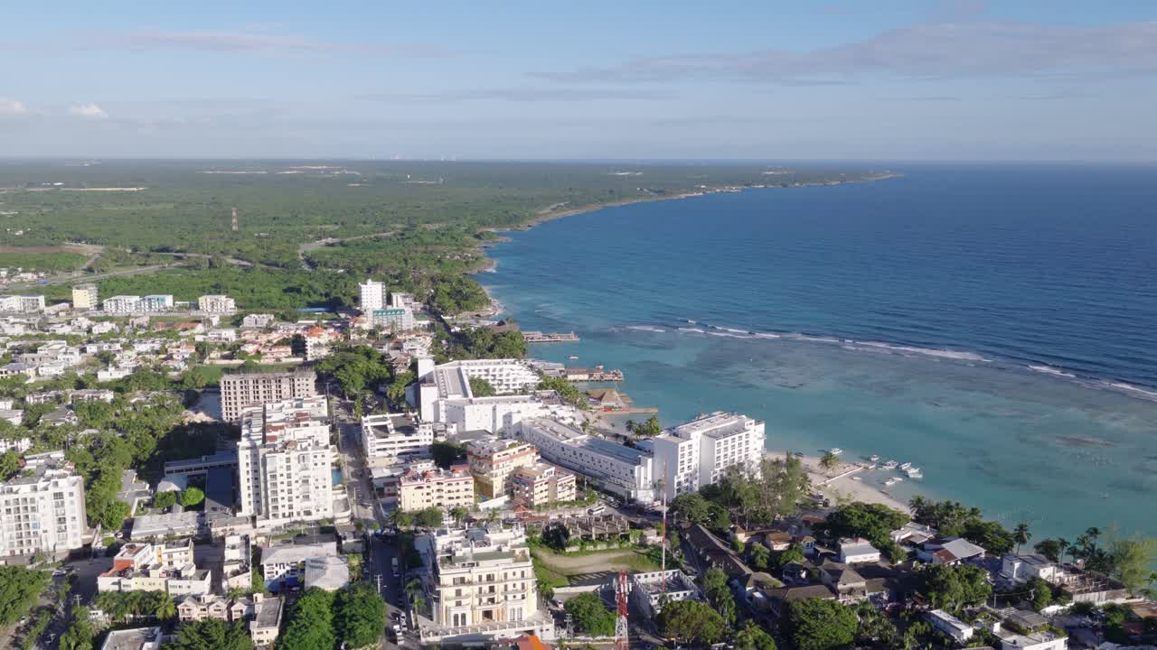 Cinematic aerial drone view of Boca Chica's cityscape and coastline. White buildings and hotels line the turquoise Caribbean Sea, Dominican Republic