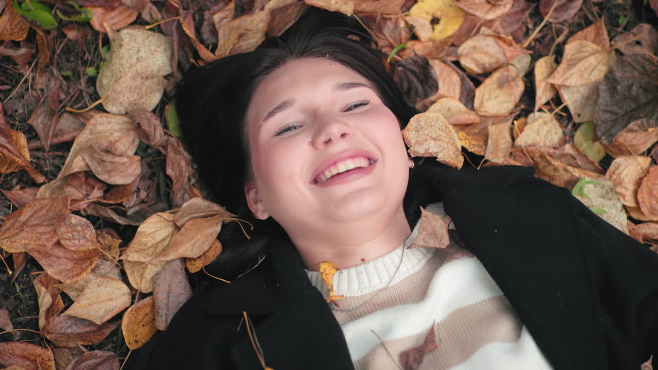 Overhead view of serene woman on forest floor making funny face among dry leaves, smiling while blocking falling leaf, cozy sweater and black coat, playful autumn mood captured with crisp texture