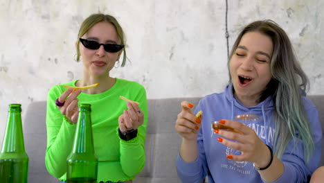 happy girl friends eating fries and having fun while sitting on sofa at home