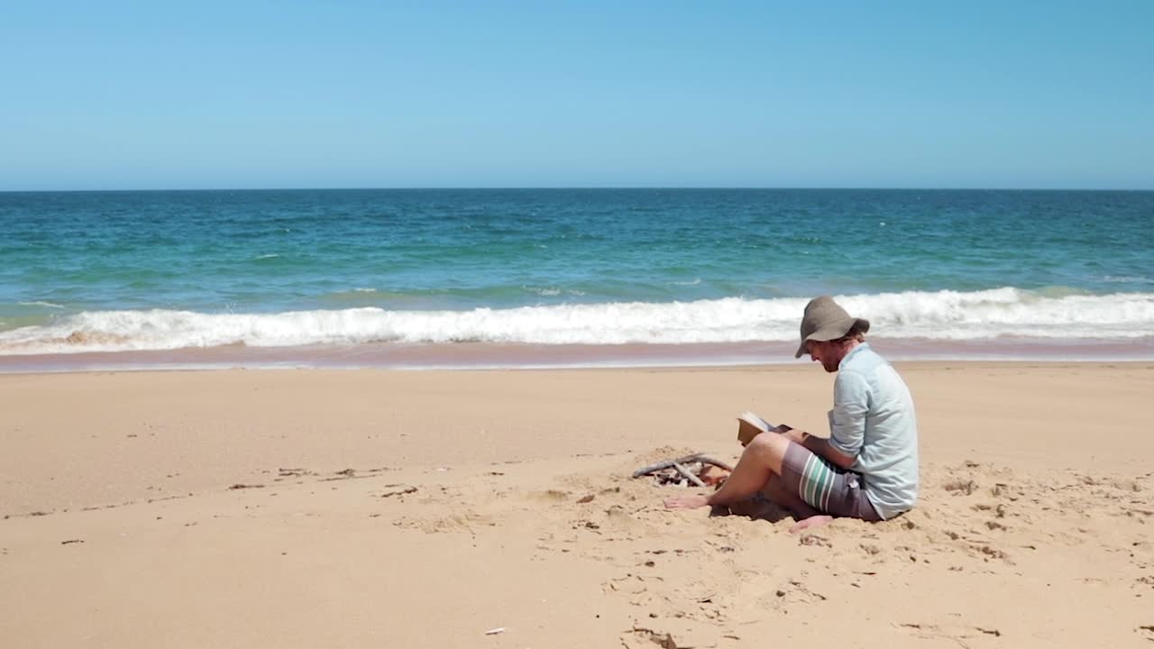 A man in a blue shirt and a hat reading a bible on the beach by a small fire.