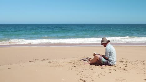 a man in a blue shirt and a hat reading a bible on the beach by a small fire.