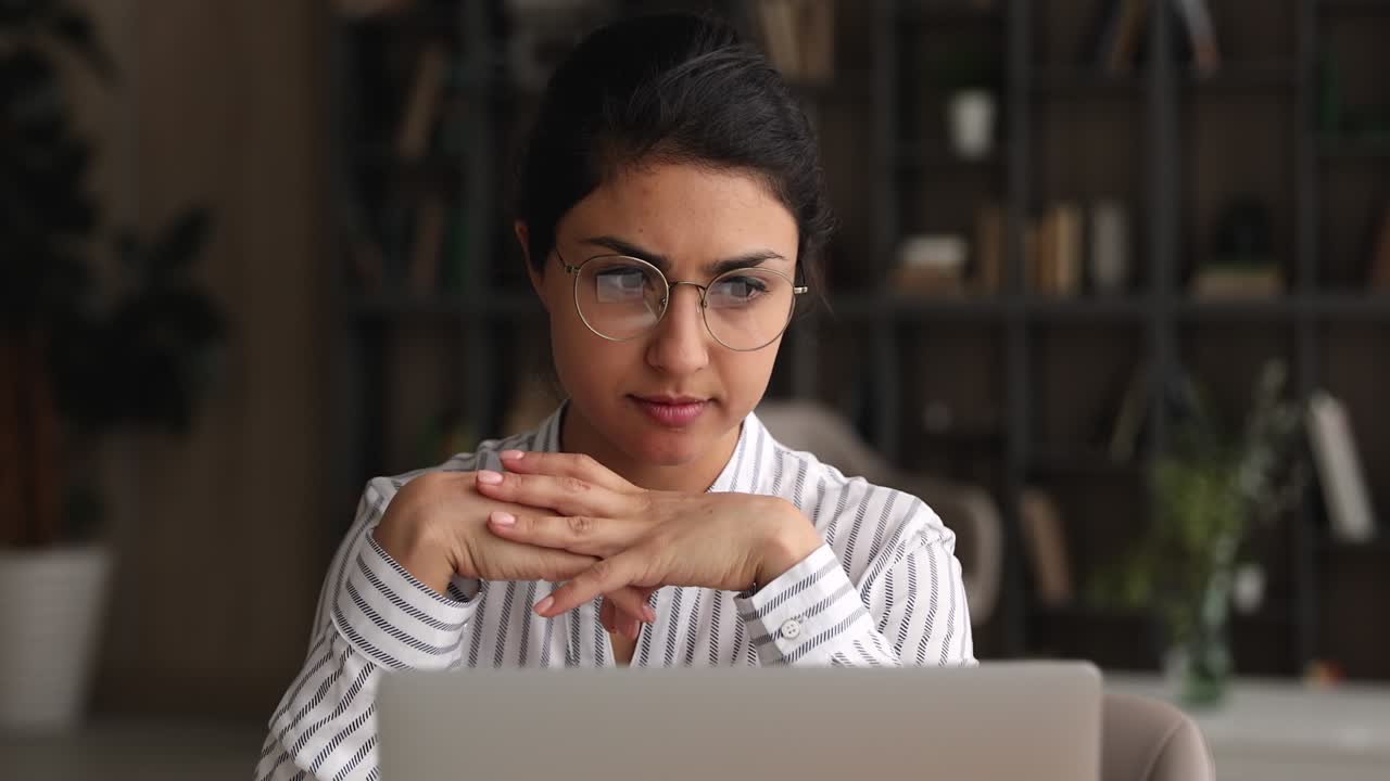 una joven empresaria india pensativa con gafas trabajando en una computadora portátil.