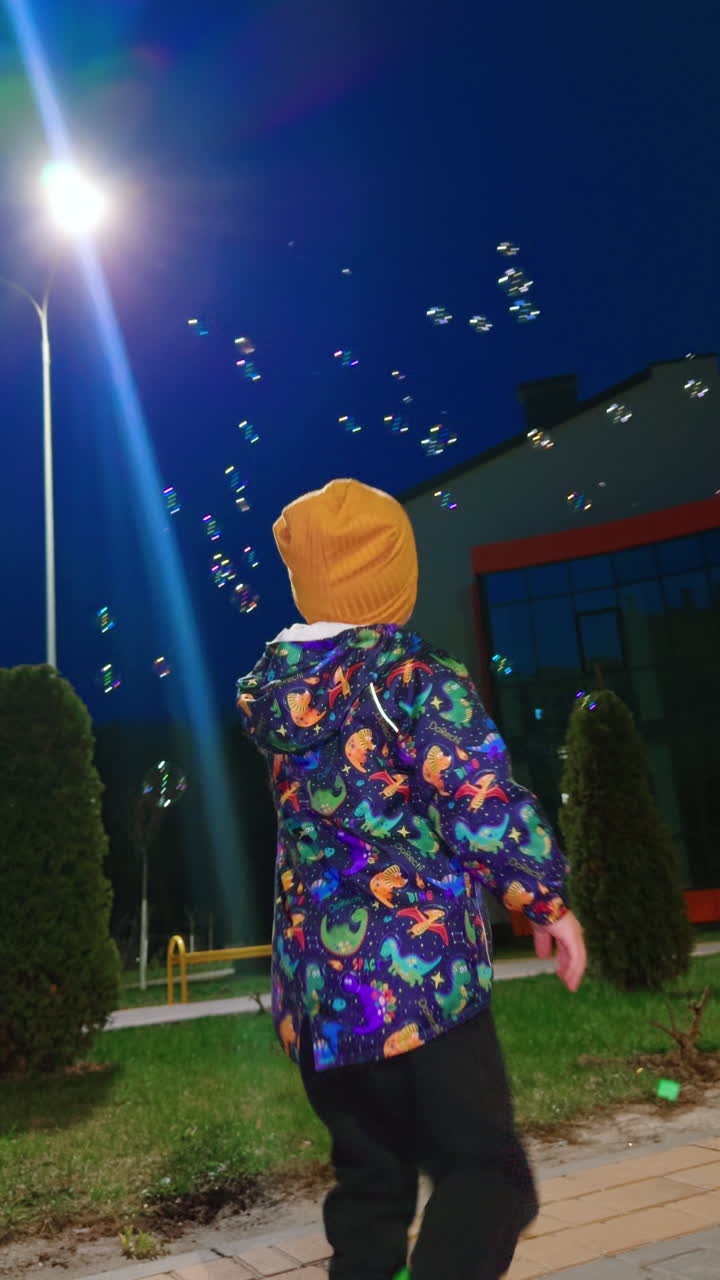 Little boy follows the soap bubbles flying around him. Low angle view on the child playing outdoors in the evening. Vertical video.