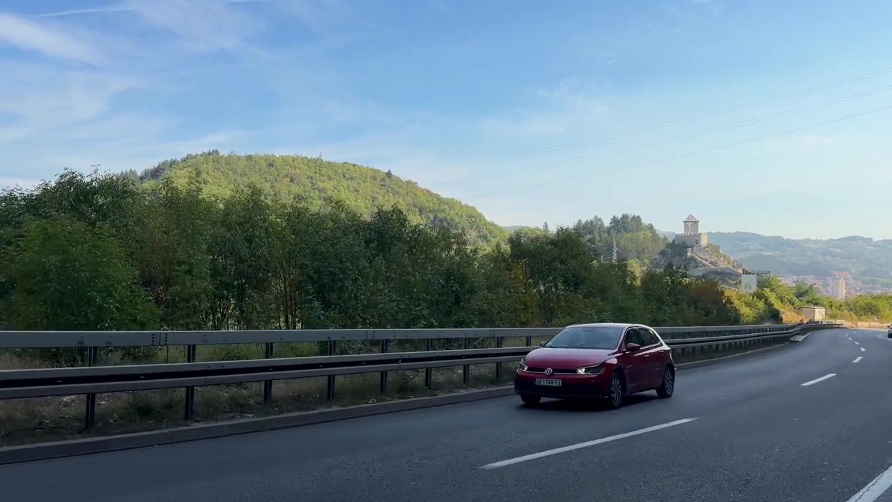 Una strada asfaltata curva attraverso verdi colline sotto la fortezza medievale di Stari Grad a Uzice, Serbia, con guardrail, alberi, montagne distanti e auto di passaggio sotto un cielo sereno