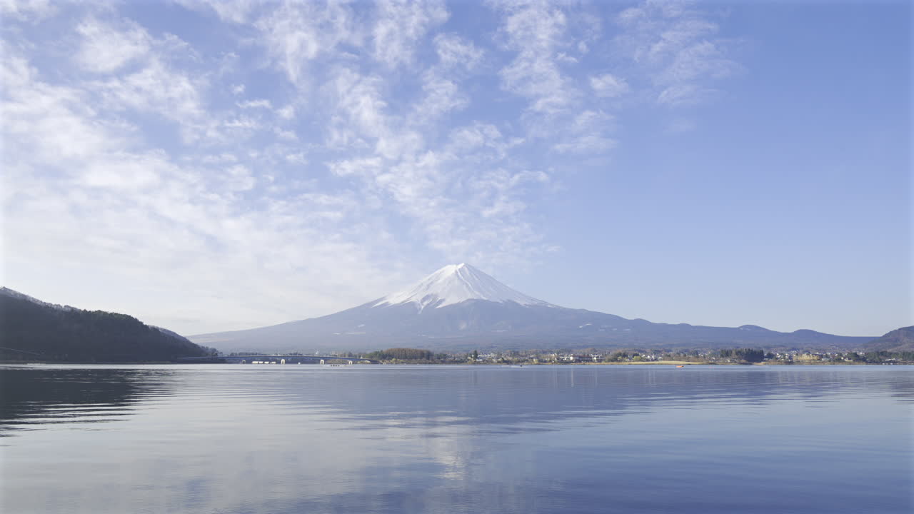 Calm waters provide a perfect reflection of Mount Fuji against a stunning sky filled with clouds. This serene location showcases the beauty of nature and offers a peaceful atmosphere. Kawaguchiko Lake