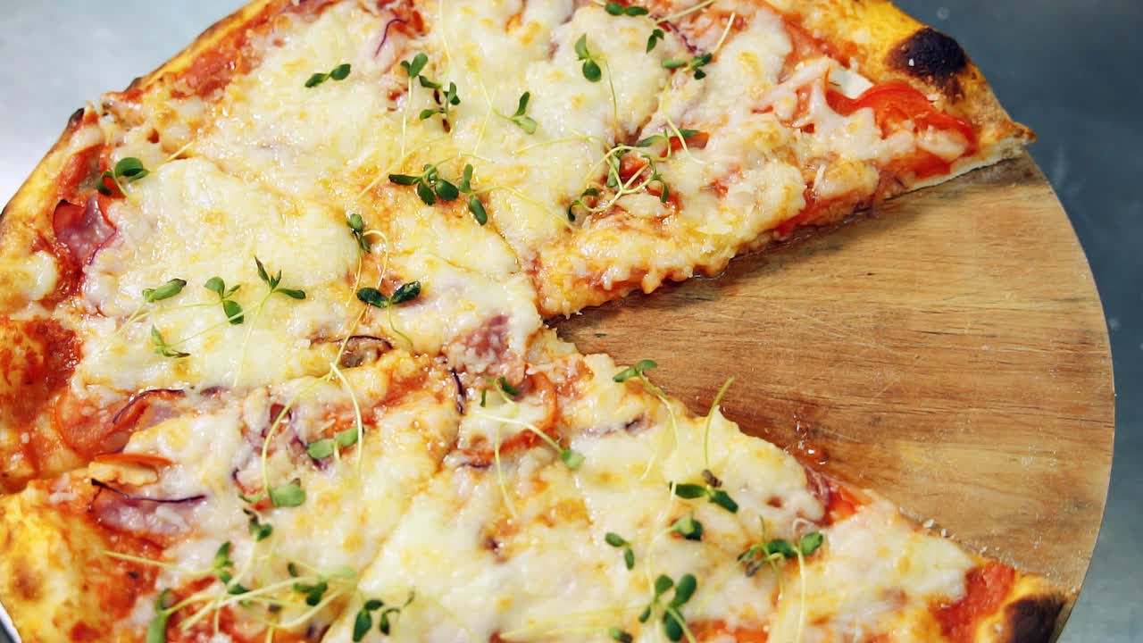 Hot fresh pizza with cheese, tomatoes and greens cut into pieces. Rotating wooden board with Italian pizza. Close-up