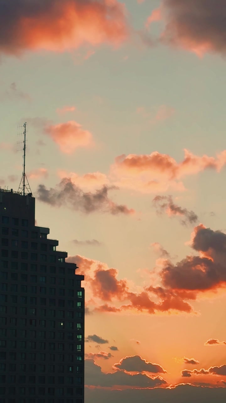 Nuvens suaves em tons de laranja e rosa flutuam sobre um céu sereno ao pôr do sol atrás de um edifício urbano em silhueta, criando uma atmosfera noturna calma.