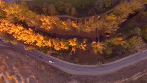 Top-Aerial-view-of-country-road-near-to-a-river-during-fall-with-beautiful-tree-colors-and-sunny-day