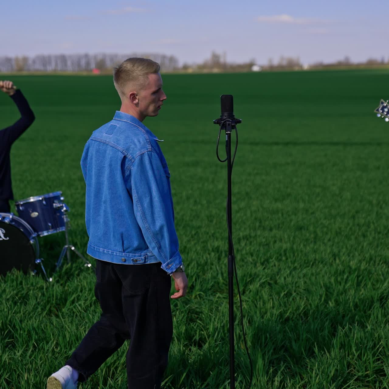 Back view of a guy in jeans jacket coming up to the microphone. Music band playing in the beautiful green field at daytime