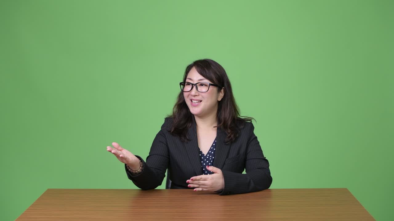 una hermosa mujer de negocios asiática madura hablando con la cámara mientras está sentada detrás del escritorio