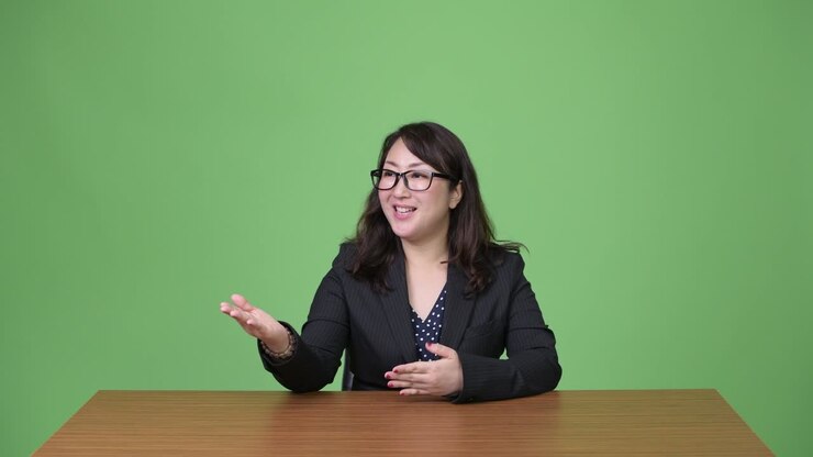Mature beautiful Asian businesswoman talking to camera while sitting behind desk