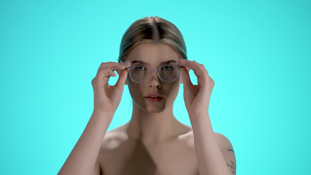 Static medium shot of a young pretty woman putting off her glasses and looking into the camera in front of turquoise background in slow motion