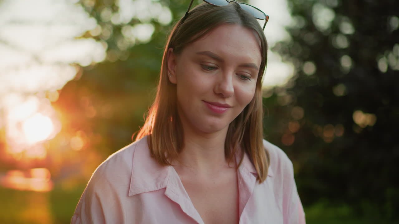 핑크 을 입고 야외에 앉아있는 아름다운 여인이 따뜻한 미소로 머리를 흔들고, 뒤로 내려다보고, 뒤에서 빛 빛은 평온하고 자연스러운 분위기를 만니다.