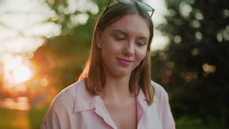 beautiful lady seated outdoors in pink top shakes head thoughtfully with a warm smile, then looks down back, sun glow from behind creates a serene, natural atmosphere