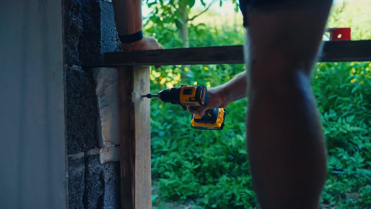 Person Using Cordless Drill To Drive Screw Into Wooden Frame Against Stone Wall. close-up shot