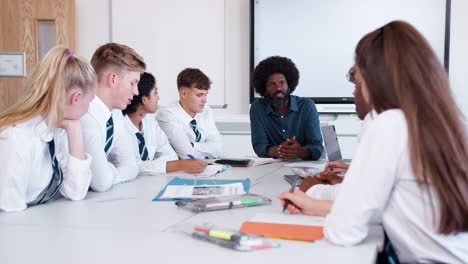 male high school teacher sitting at table with teenage pupils wearing uniform teaching lesson