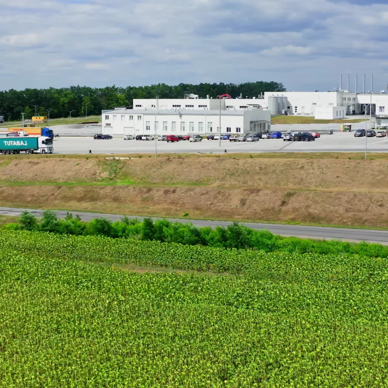 Agricultural land near the modern manufacturing. Green field near the road. Large factory in the rural place. Truck moving near the industrial zone