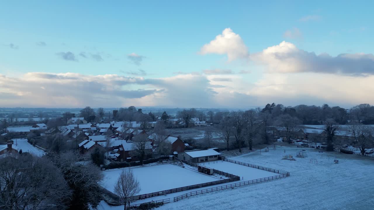 A drone shot captures a peaceful English village, its snow-covered rooftops glowing under the bright winter sun.