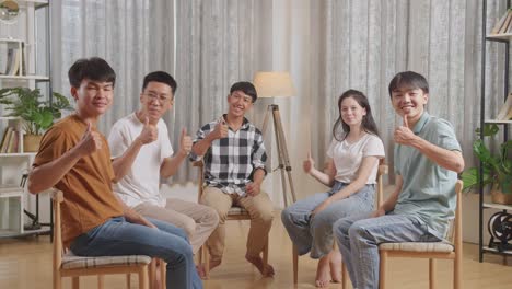 asian teen group sitting in chairs forming a circle discussing at home. smiling and showing thumbs up gesture to camera