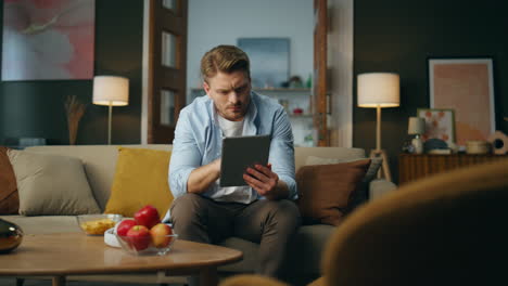 Pensive freelancer swiping tablet cozy interior. Serious guy sitting sofa alone