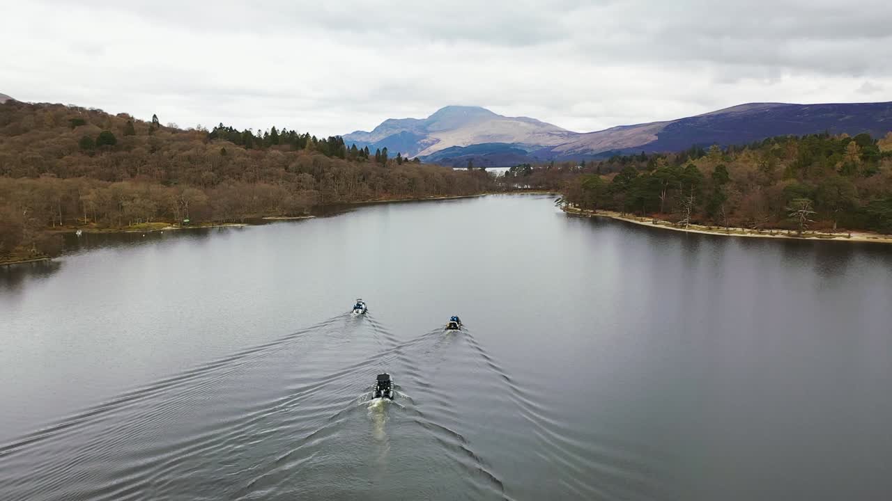 Cinematic slowmo aerial view of a flyby of 3 boats approaching the narrows on Loch Lomond in Scotland, lifting up to reveal Ben Lomond