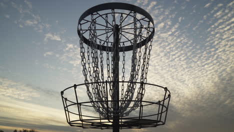 disc golf basket silhouetted by evening sky while disc golfer makes and retrieves putt made with a distance driver