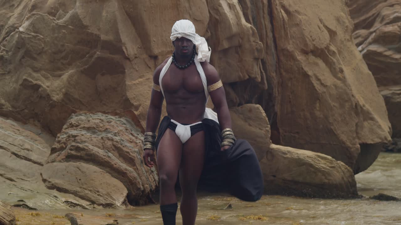 A dark-skinned, muscular male cosplayer poses on the beach.