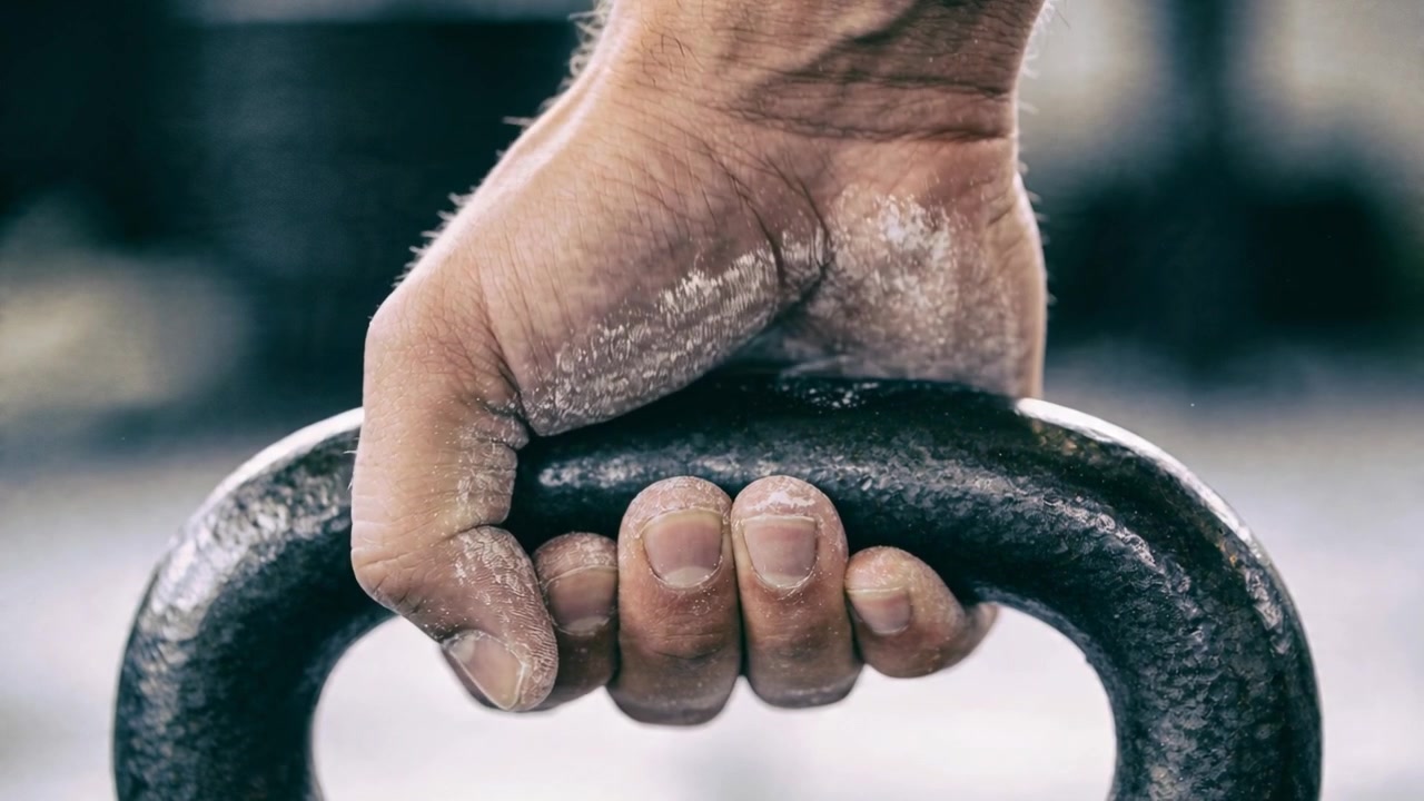Mão adulta ajustando a pegada no cabo do kettlebell na academia, preparando-se para o levantamento com marcas de giz.
