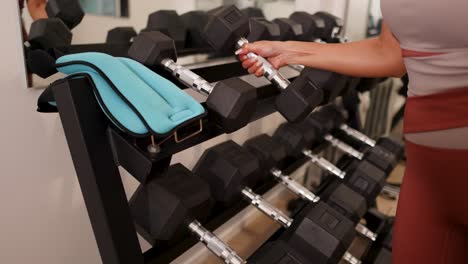 a woman chooses and lifts dumbbells from a rack in a well-lit gym setting, showcasing fitness and strength