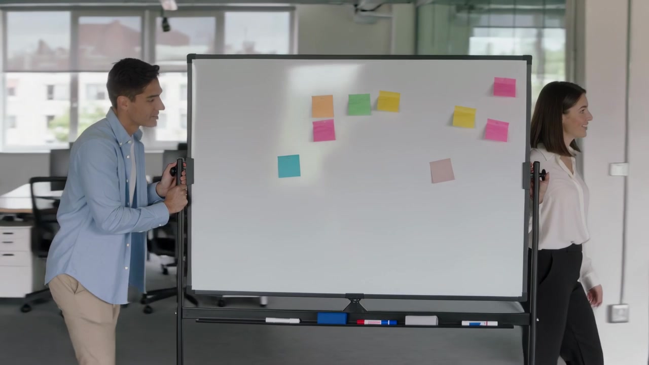 Kokous, jossa henkilökunta työntää valkotaulua toimiston poikki; mies sinisessä paidassa, nainen valkoisessa puserossa, post-it-laput