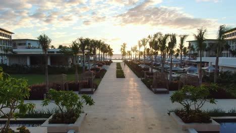 Luxury-all-inclusive-resort-at-sunset,-modern-pool-area-with-lounges-and-broad-alley,-tropical-beach-with-palm-trees-in-the-background,-dramatic-cloudscape,-aerial-dolly-view