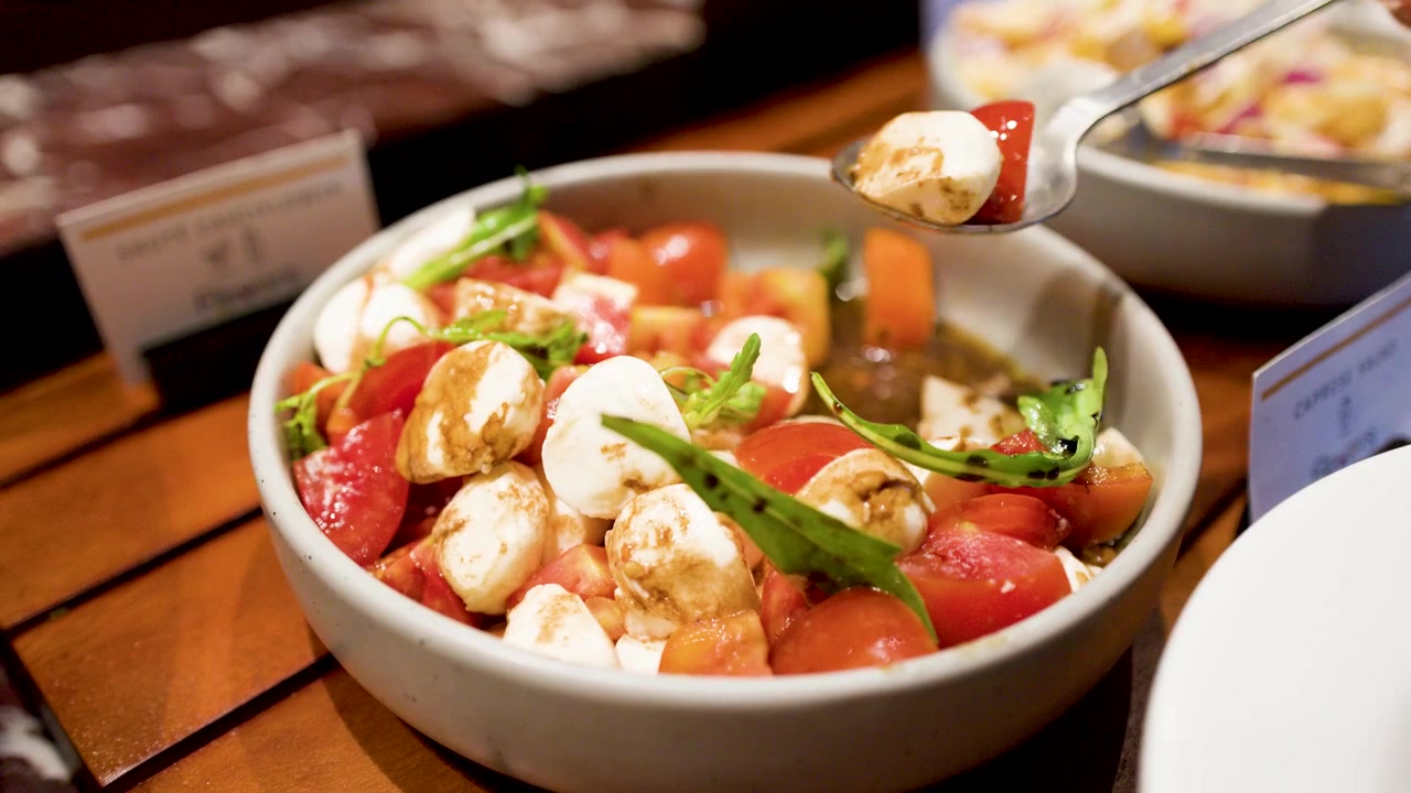 A metal spoon scoops fresh mozzarella and tomatoes from a bowl at an international buffet