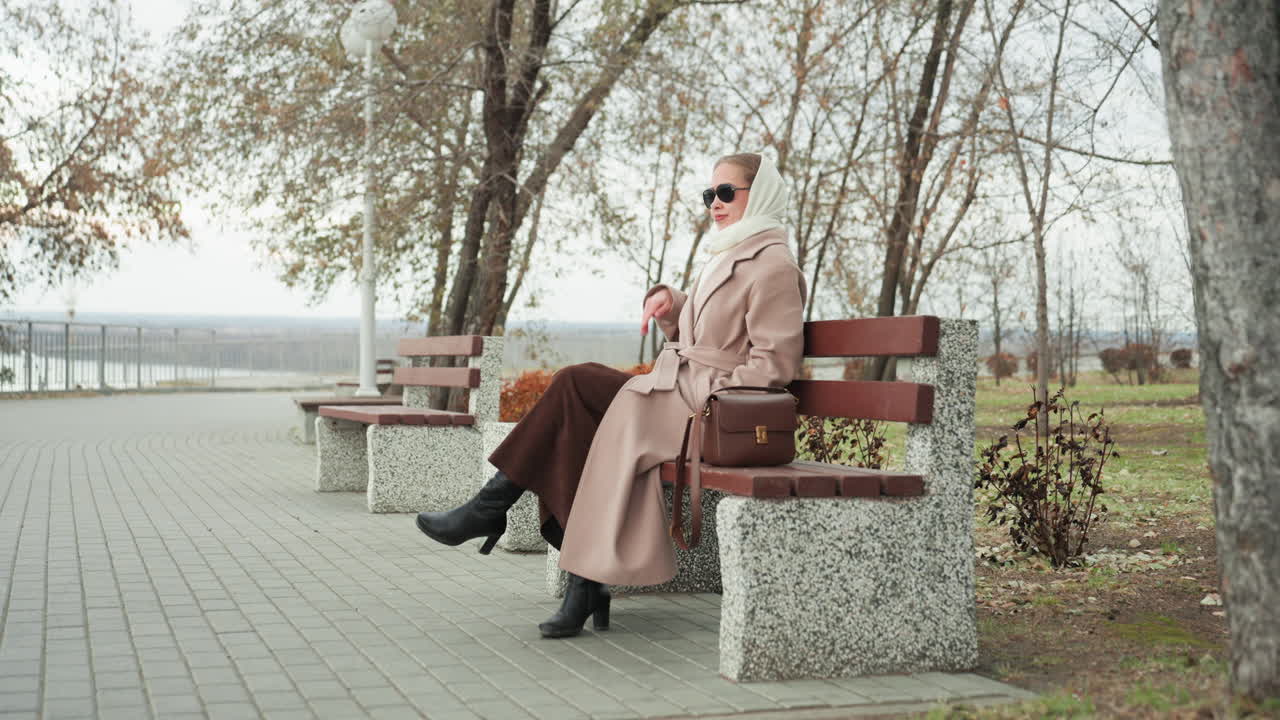 Donna elegante che sorseggia caffè su una panchina, con occhiali da sole e cappotto lungo, borsa in pelle sul sedile, momenti con lo smartphone, in un parco sul lungomare con l'aria frizzante d'autunno, postura sicura e rilassata, con un'atmosfera da editoriale di moda.