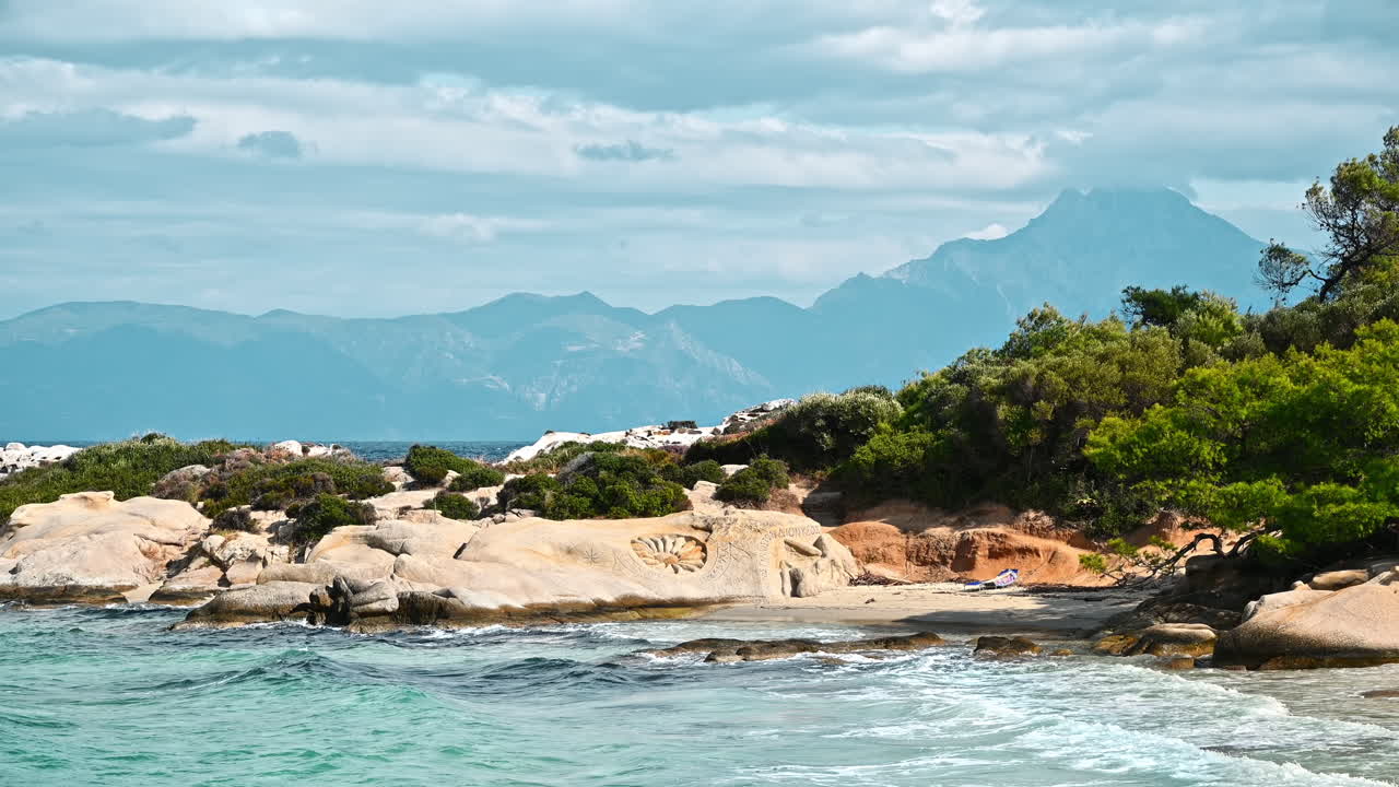 Aegean sea coast with greenery around, rocks, bushes and trees, blue water with waves, Greece